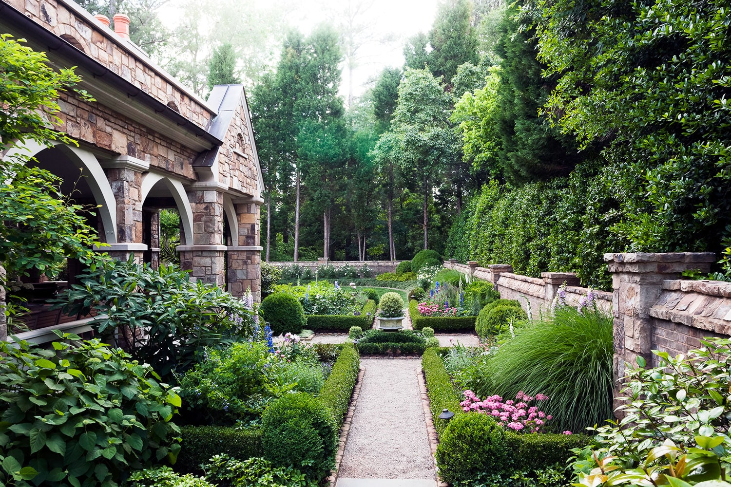 A formal garden beside a stone house, featuring manicured hedges, flowering plants, topiary shrubs, and a gravel pathway bordered by green lawns and vibrant flowers. Tall trees and stone walls enclose the tranquil garden area.