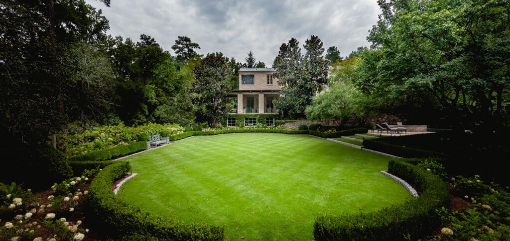 A large, manicured lawn bordered by symmetrical hedges and flowerbeds leads to a two-story brick house surrounded by tall trees under a cloudy sky. Benches and pathways are arranged neatly in the garden.