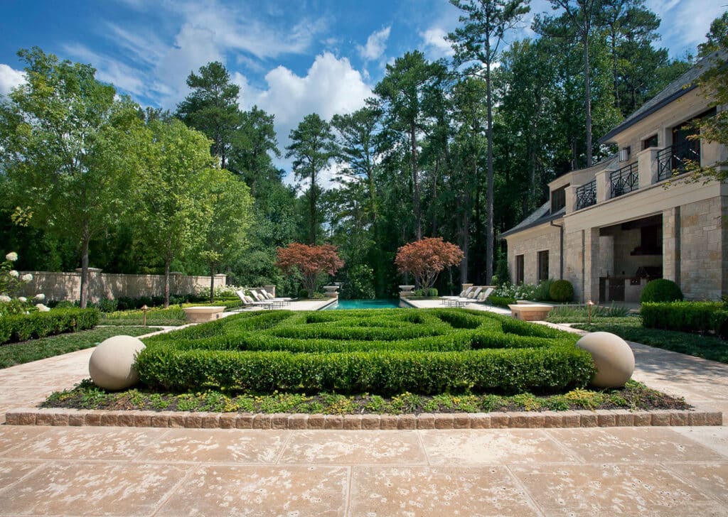 Elegant landscaped backyard with manicured boxwood hedges, stone pathways, decorative spheres, lounge chairs, a swimming pool, and a large house, surrounded by tall trees under a blue sky with clouds.
