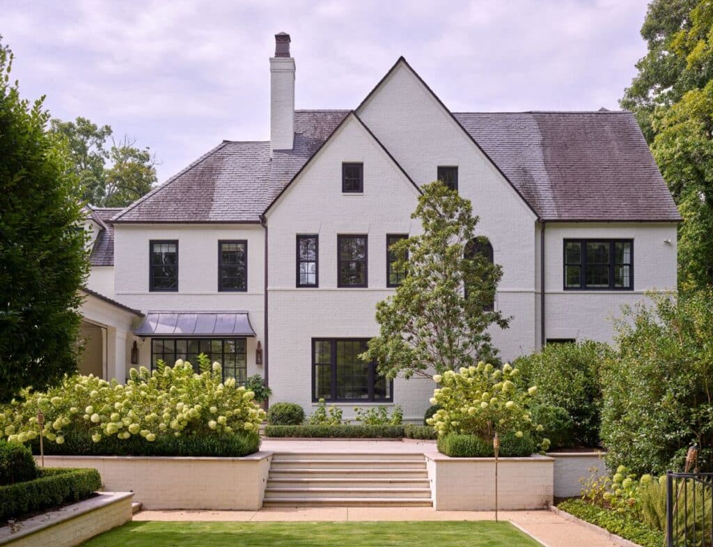 A large, two-story white house with black-framed windows, a steep roof, and a central chimney. The home is surrounded by manicured gardens, bushes, hydrangeas, trees, and a wide set of steps leading to the entrance.