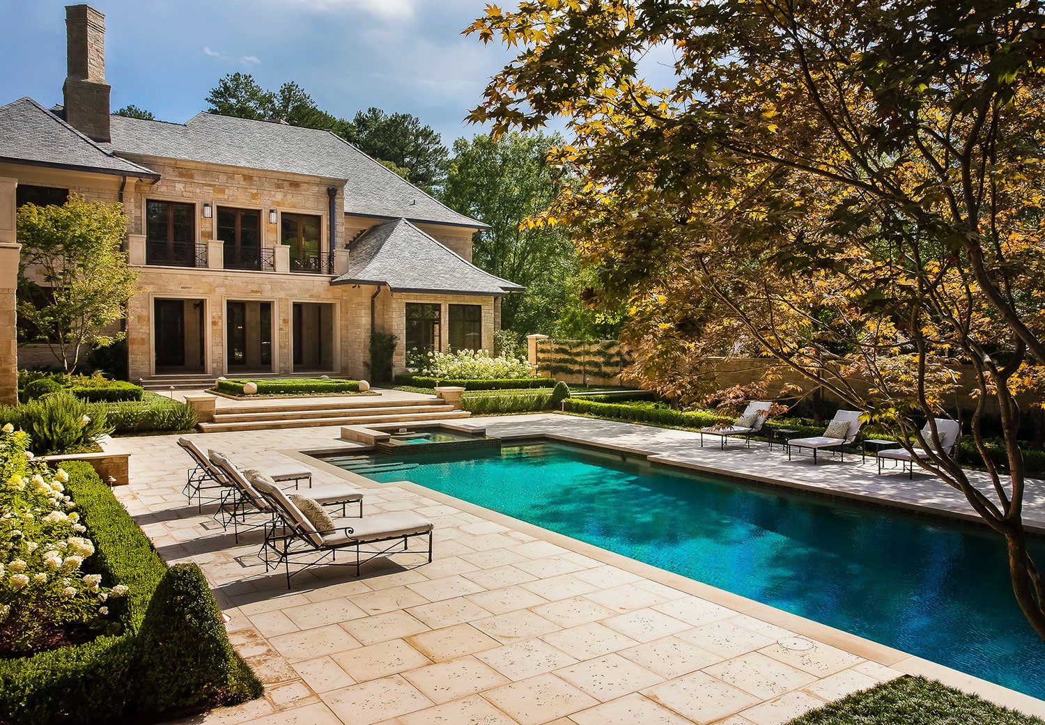 Elegant stone mansion with large windows overlooks a rectangular swimming pool surrounded by a paved patio, lounge chairs, manicured bushes, and trees under a blue sky.