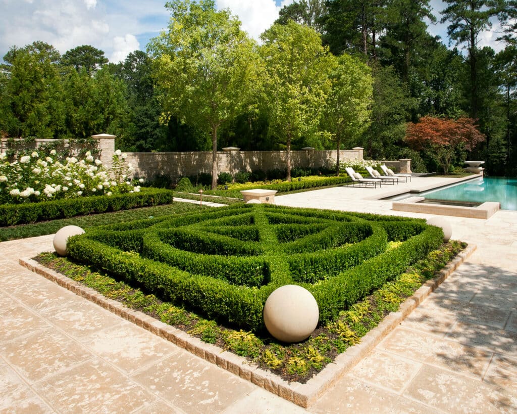 A formal garden with geometric boxwood hedges, stone spheres at the corners, manicured lawns, flowering bushes, lounge chairs, and a modern rectangular swimming pool surrounded by trees.
