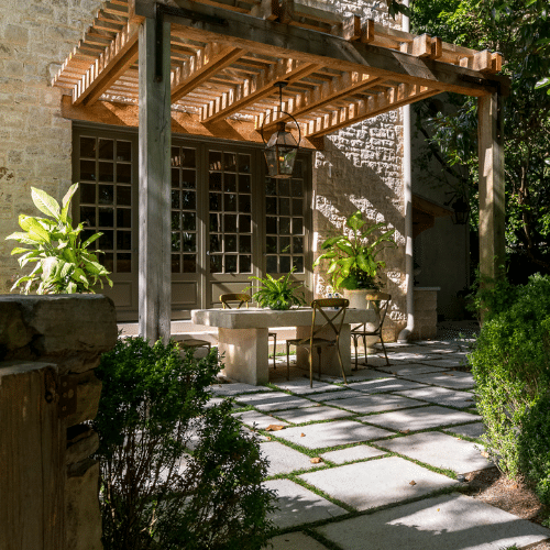 Large cement table on paved area under pergola with ferns and other plants along border