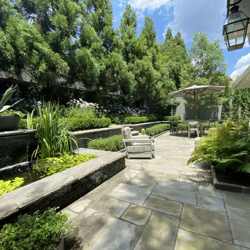 Paved terrace with seating area, large trees for privacy, and fountain with water plants.