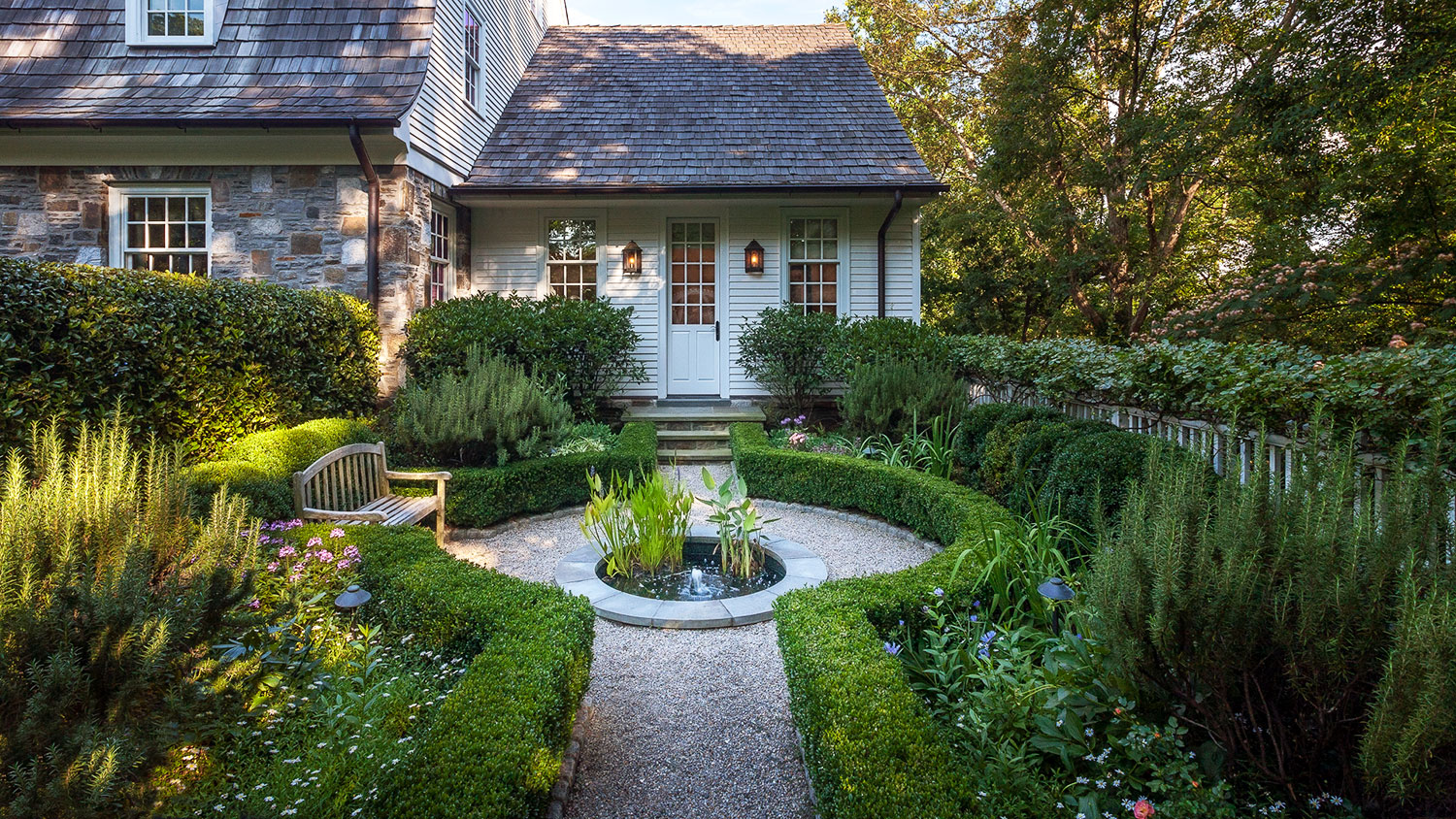 A beautiful pea gravel path bordered by korean boxwood hedges and blooming garden that continues to a New England style home.